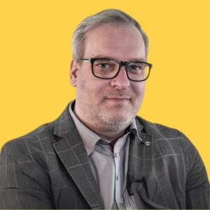 Mężczyzna w okularach w czarnej oprawce i szarej marynarce w kratę, uśmiecha się na żółtym tle.
