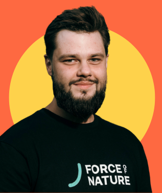 Young man with a full beard wearing a black shirt with "FORCE OF NATURE" logo, against a yellow and orange background.