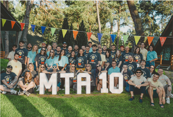 Large group outdoors posing behind white 'MTA 10' letters, many wear matching shirts.
