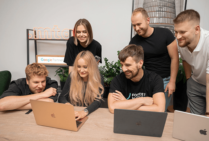 Five smiling colleagues collaborate around a wooden table, looking at a rose gold laptop screen.