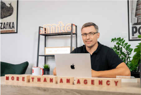 Man smiling at desk with MacBook, in front of 'More Than An Agency' blocks.