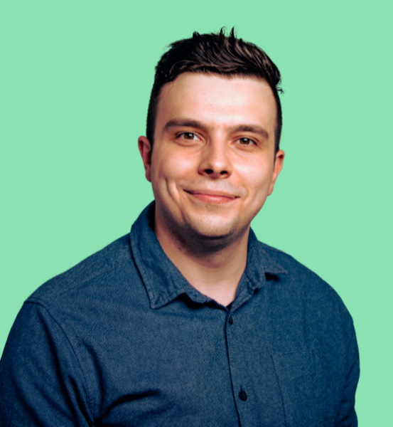 Mężczyzna o ciemnych włosach, ubrany w granatową koszulę, uśmiecha się ciepło na jasnoróżowym tle.