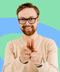 Portret młodego mężczyzny w okularach, z brodą i jasnymi włosami, ubranego w beżowy sweter. Uśmiechnięty, spogląda prosto w obiektyw i wskazuje palcami w stronę aparatu. Tło w odcieniach zieleni i błękitu dodaje zdjęciu nowoczesnego, energetycznego charakteru.