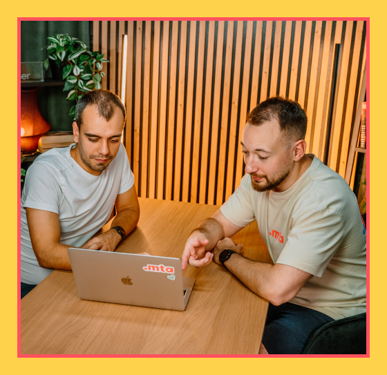 Grafika przedstawia dwóch mężczyzn pracujących wspólnie przy laptopie z logo MTA Digital. Obaj skupieni są na ekranie – jeden z nich wskazuje coś palcem, drugi uważnie się przygląda. Scena rozgrywa się w nowoczesnym, przytulnym biurze z drewnianą lamelową ścianą i zielenią w tle.