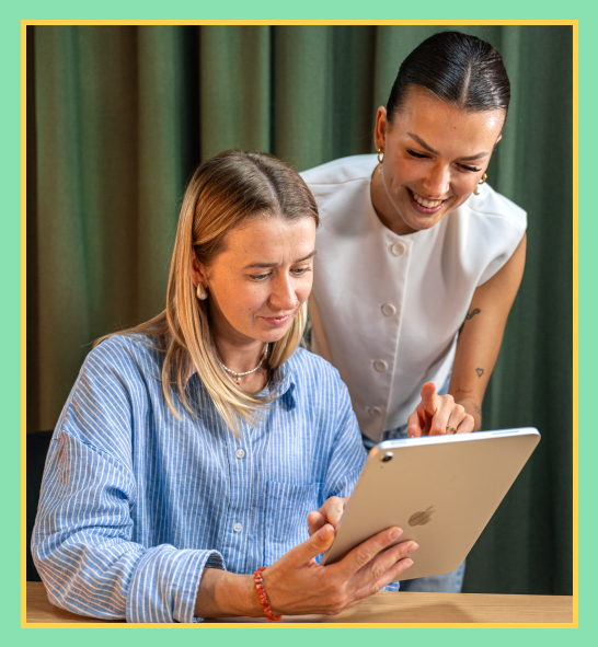 Grafika przedstawia dwie kobiety zaangażowane we wspólną pracę przy tablecie. Jedna z nich siedzi i trzyma urządzenie w dłoniach, druga pochyla się z uśmiechem, wskazując coś na ekranie.