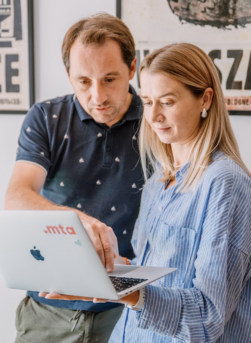 Zdjęcie przedstawia dwie osoby z zespołu MTA Digital pracujące wspólnie przy laptopie. Mężczyzna w granatowej koszulce wskazuje coś na ekranie, a kobieta w niebieskiej koszuli uważnie się przygląda. Na obudowie laptopa widoczne jest czerwone logo .mta i serduszko. Tło stanowią plakaty w ramkach, które nadają wnętrzu charakteru. Kadrowanie i naturalne światło budują atmosferę zaangażowanej współpracy.