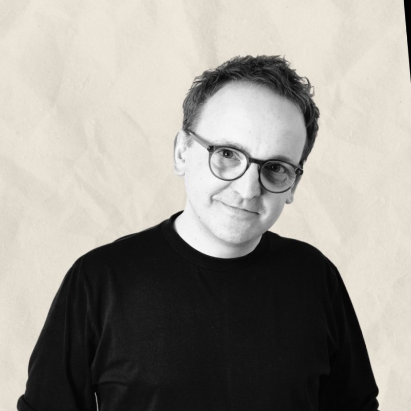 Grafika przedstawia portret Sebastiana Śniecińskiego w czarno-białej stylistyce. Postać stoi lekko bokiem, skierowana w stronę obiektywu, z subtelnym, spokojnym uśmiechem. Ma krótkie włosy oraz nosi okrągłe okulary. Ubrany jest w prostą, ciemną koszulkę, co w połączeniu z neutralnym, jasnym tłem nadaje zdjęciu minimalistyczny i wyważony charakter. Całość buduje wizerunek ekspercki, spokojny i skupiony na osobie.