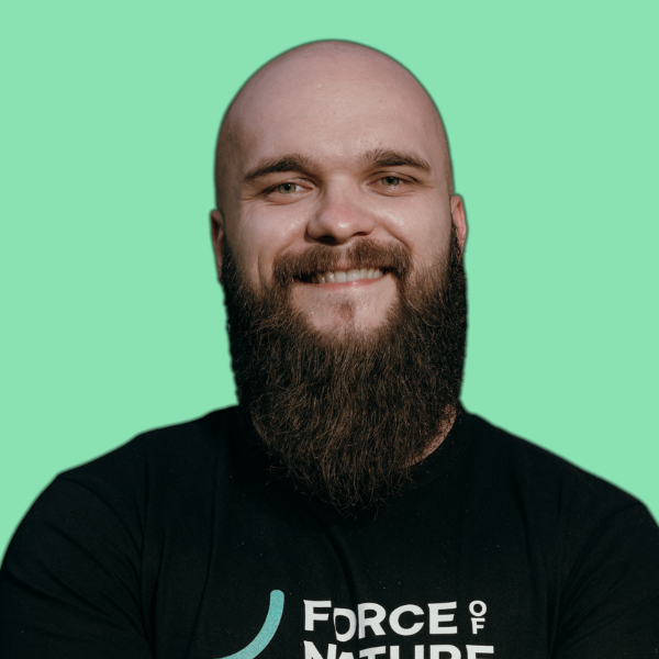 Zdjęcie przedstawia Jacka Matuszewskiego, który stoi przodem do obiektywu i uśmiecha się. Ma ogoloną głowę, długą brodę oraz ubrany jest w czarną koszulkę z nadrukiem. Tło jest jednolite, w odcieniu zieleni, co podkreśla minimalistyczny i nowoczesny charakter fotografii. Całość ma profesjonalny, a jednocześnie swobodny wydźwięk, wpisujący się w komunikację zespołu MTA Digital.