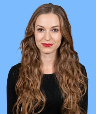 Portrait of a young woman with long, wavy brown hair and red lips, against a light blue background.