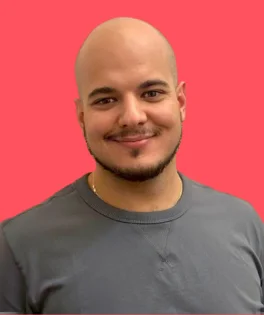 Young, bald man with short stubble, smiling, wearing a grey sweatshirt, against a light pink background.