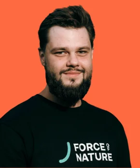 Young man with a beard, wearing a black T-shirt with a 'FORCE OF NATURE' logo, against an orange background.