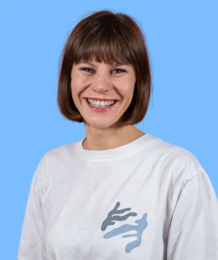 Smiling woman with short brown hair and bangs, wearing a white T-shirt with a blue print, against a light blue background.
