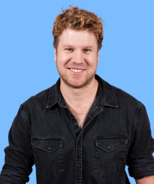 Smiling, fair-haired man wearing a black denim shirt, against a light blue background.