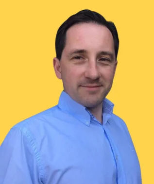 Man with dark hair, wearing a light blue collared shirt, smiling against a bright yellow background.