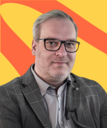 Błażej Cybowski – mężczyzna w okularach i szarej marynarce w kratę, na tle żółto-czerwonej grafiki