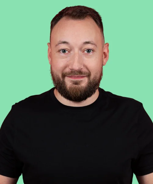 Man with short hair and a full brown beard, wearing a black T-shirt, smiling against a light turquoise background.
