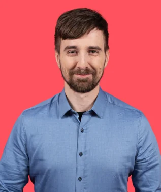 Man with a beard and short hair, wearing a blue shirt, smiling against a bright pink background.